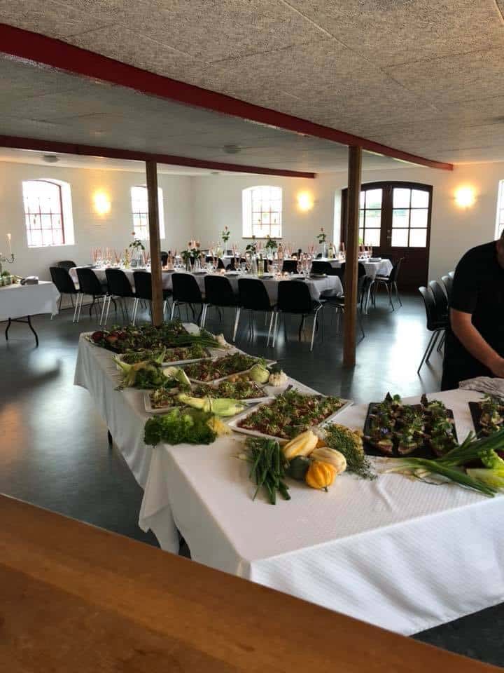 Buffetbord med anretninger og grønt i forgrunden og festsalen dækket op med langborde, vinglas og lys i baggrunden